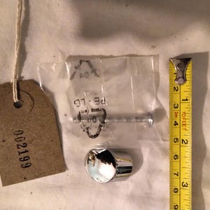 May include: An assortment of hardware components, including a chrome-finished cap, a bolt in a plastic bag, and a brown tag with the number "002199". A yellow measuring tape is also present, showing measurements in inches.