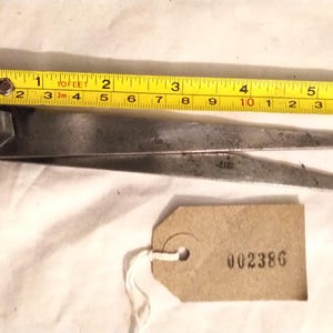 May include: A metal tool with pointed ends, possibly for a specific trade or craft. A yellow tape measure is placed above the tool, showing measurements in inches. A tag with the number "002386" is attached.