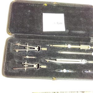 May include: A black rectangular case containing a set of silver-toned metal drafting instruments, including two compasses, a divider, and various attachments. The interior is lined with black velvet, and a white rectangular label is attached.