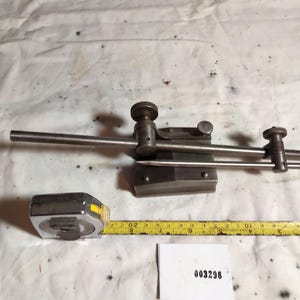 May include: A grey metal surface gauge tool featuring a heavy base, a vertical post, and an adjustable scriber arm with two knurled adjustment knobs. A yellow measuring tape shows the tool is approximately 10 inches long. A white tag displays 003296.