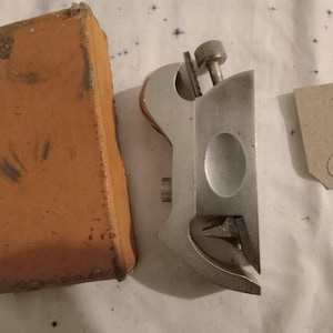 Peut inclure: Un outil métallique ancien de couleur argentée, avec une vis et un design incurvé. Il est accompagné d'une boîte en carton orange usée et d'une petite étiquette avec le numéro "002524". L'outil semble être vintage.