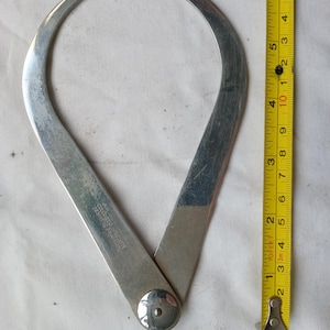 May include: A vintage silver-colored outside caliper tool, open and positioned next to a yellow measuring tape. The caliper is hinged at the bottom and has curved arms. The measuring tape shows inches.