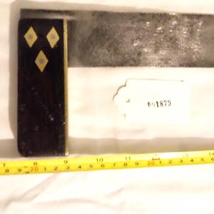 May include: A vintage metal try square with a black handle featuring three gold diamond-shaped inlays. A yellow measuring tape is next to the square, showing measurements in inches. A tag with the number "01879" is also present.