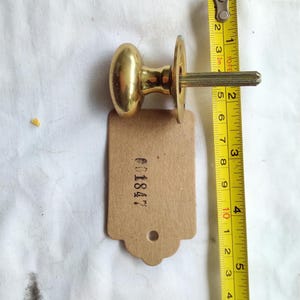 Könnte beinhalten: Ein Messing-Türknauf mit passender Rückplatte und Spindel, mit einem braunen Anhänger mit der Aufschrift "001847". Der Knauf misst etwa 5 cm in der Länge. Ein gelbes Maßband liegt neben dem Knauf.