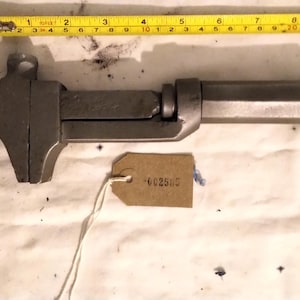 May include: A vintage adjustable wrench, approximately 25 cm long, with a dark grey finish. A yellow measuring tape is placed above the wrench. A small brown tag with the number "002585" is attached to the wrench.