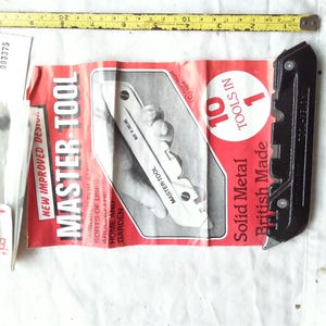 May include: A black metal utility knife positioned next to a red and white product instruction sheet labeled Master-Tool 10 Tools in 1. A yellow measuring tape at the top shows the tool length is approximately 15 cm. A small plastic bag is included.