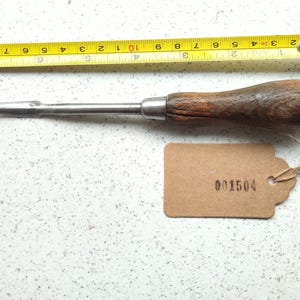 May include: A vintage wood-handled metal tool with a pointed tip. The tool is about 15 cm long and has a brown wooden handle. A brown paper tag with the number "001504" is attached to the tool.