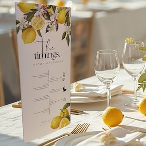 May include: A table setting with a lemon theme, featuring a floral arrangement of yellow roses and greenery. Fresh lemons are placed on the white tablecloth. Wine glasses, plates, and gold cutlery create an elegant look. A menu with the text "The timings" is visible.