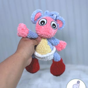 May include: A crocheted plush toy in the shape of a small animal with a pink body, blue ears, and red shoes. The toy is wearing a white diaper and has large black eyes.
