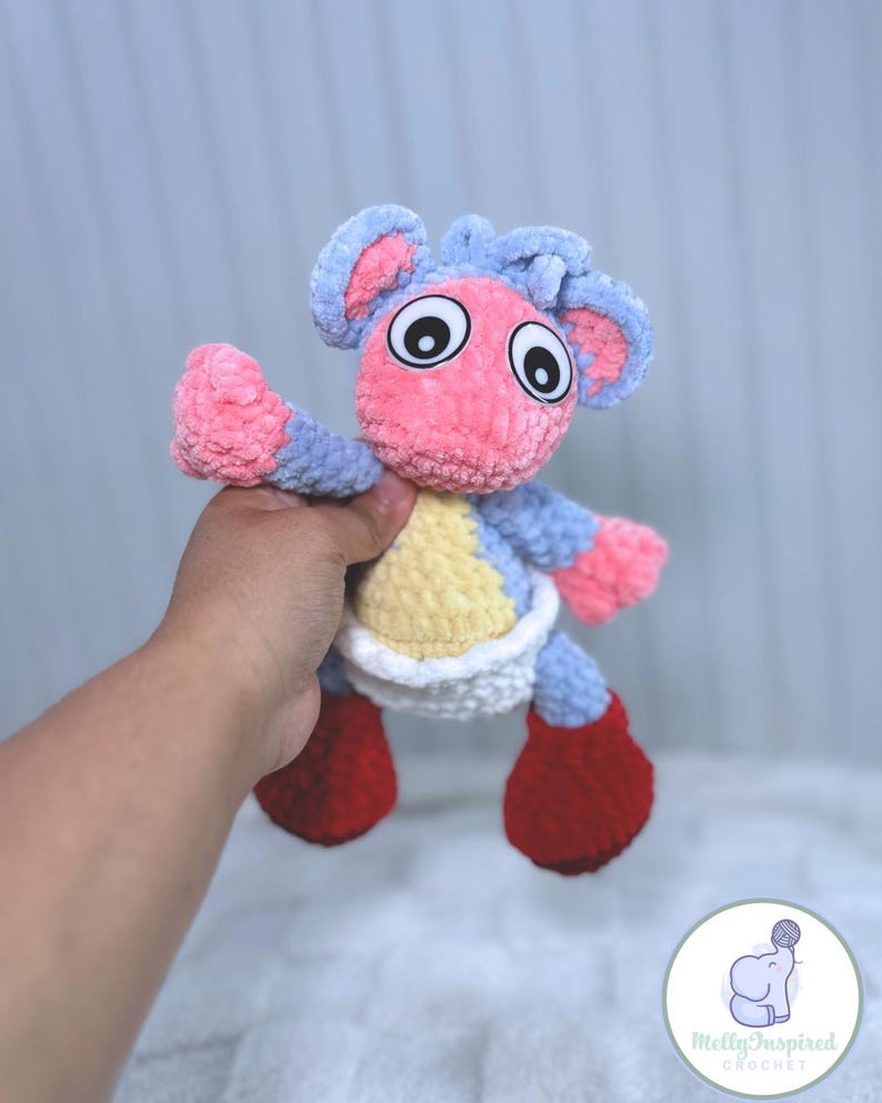 May include: A crocheted plush toy in the shape of a small animal with a pink body, blue ears, and red shoes. The toy is wearing a white diaper and has large black eyes.