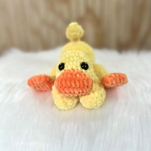 May include: A crocheted yellow duck toy with orange beak and feet. The duck is lying on its belly with its head facing forward.