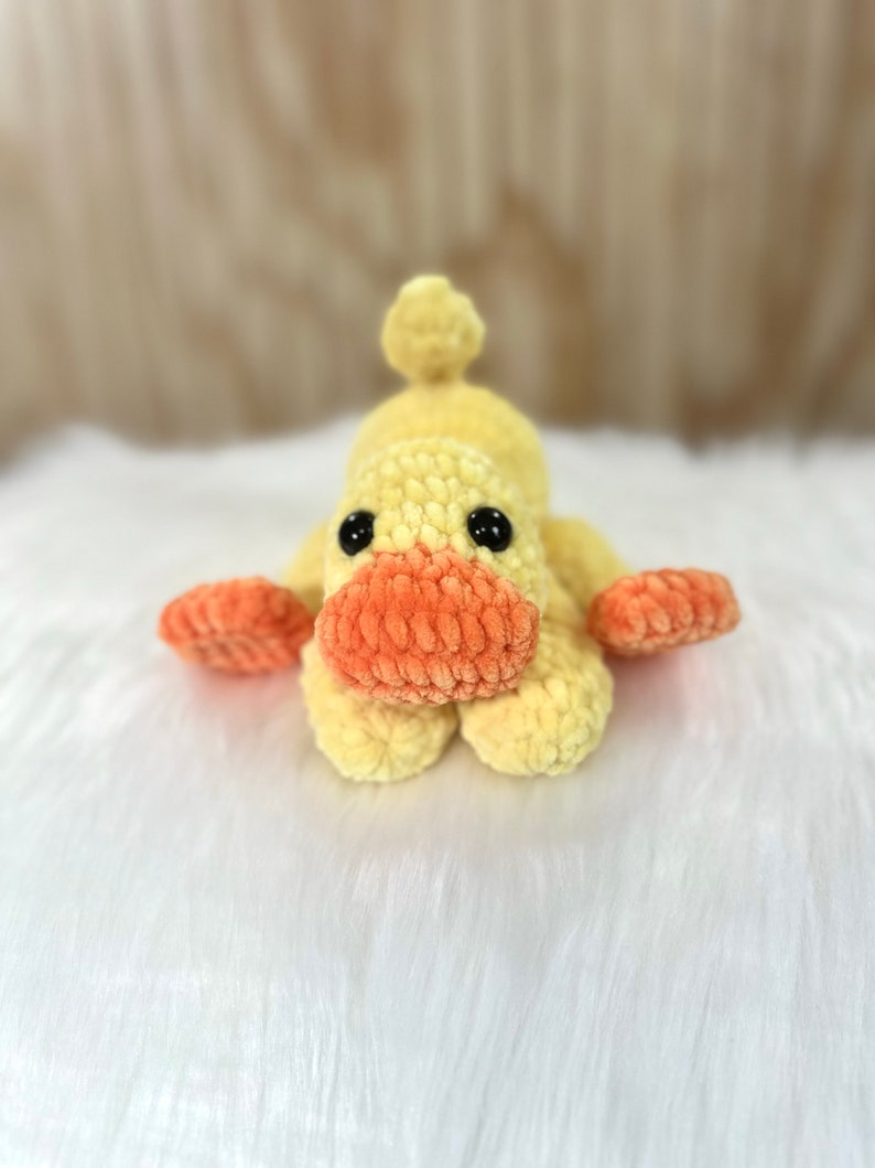 May include: A crocheted yellow duck toy with orange beak and feet. The duck is lying on its belly with its head facing forward.