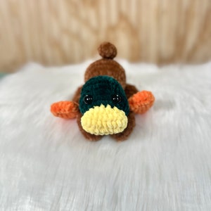 May include: A crocheted duck plush toy with brown, green, orange, and yellow yarn. The duck has black eyes and a yellow beak.