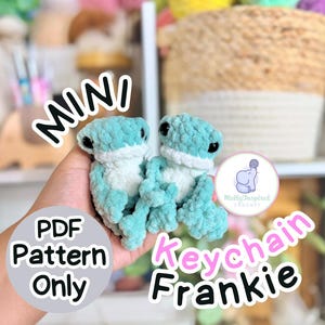 May include: Two turquoise and white crochet frog keychains. The keychains are labeled "Keychain Frankie" and "PDF Pattern Only". The frogs have black eyes and are made of a soft, textured yarn. The background is blurred, with yarn and a basket visible.