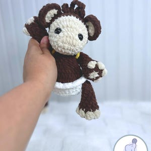 May include: A brown crocheted monkey toy wearing a white diaper. The monkey has a yellow yarn detail around its neck and brown yarn hair. The toy is being held by a hand.