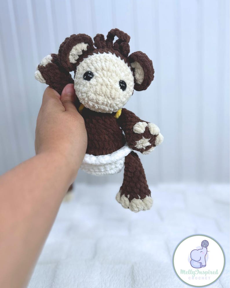 May include: A brown crocheted monkey toy wearing a white diaper. The monkey has a yellow yarn detail around its neck and brown yarn hair. The toy is being held by a hand.