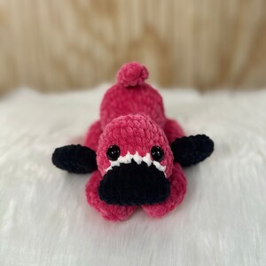 May include: A pink crocheted platypus stuffed animal with black eyes and a black crocheted beak. The platypus is lying on a white fluffy surface.