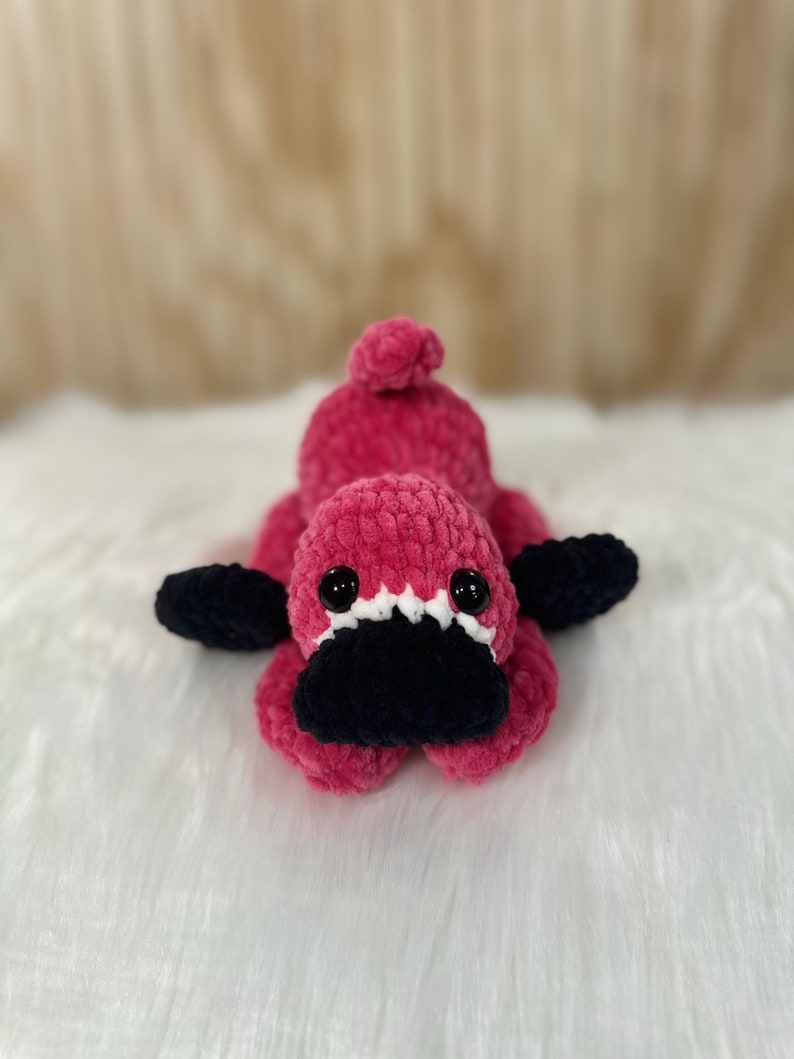 May include: A pink crocheted platypus stuffed animal with black eyes and a black crocheted beak. The platypus is lying on a white fluffy surface.