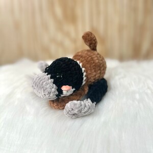 May include: A crocheted plush toy shaped like a platypus. The toy is brown, black, and gray with a pink nose and black eyes. The toy is lying on a white fluffy surface.