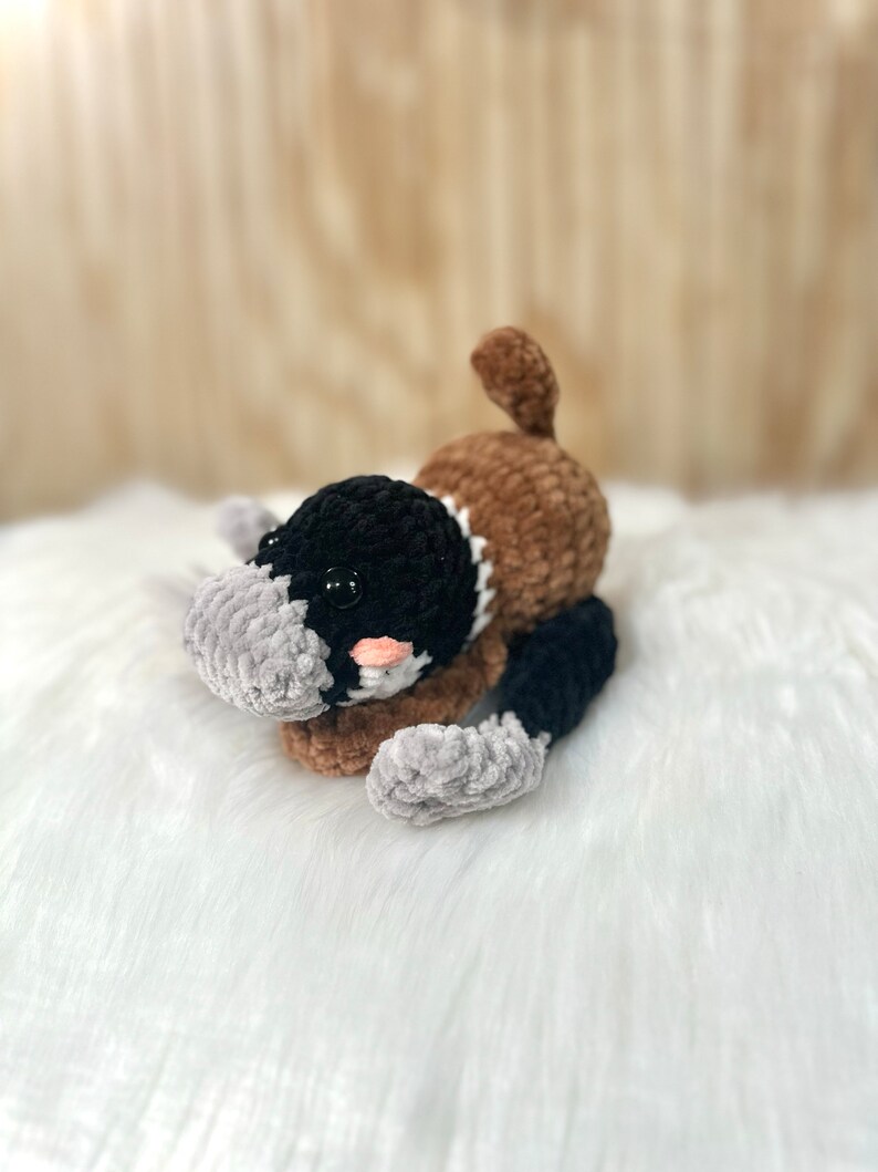May include: A crocheted plush toy shaped like a platypus. The toy is brown, black, and gray with a pink nose and black eyes. The toy is lying on a white fluffy surface.