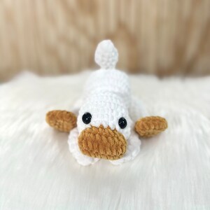 May include: A white crocheted duck plush toy with brown crocheted eyes and a brown crocheted beak. The duck is lying on a white fluffy surface.