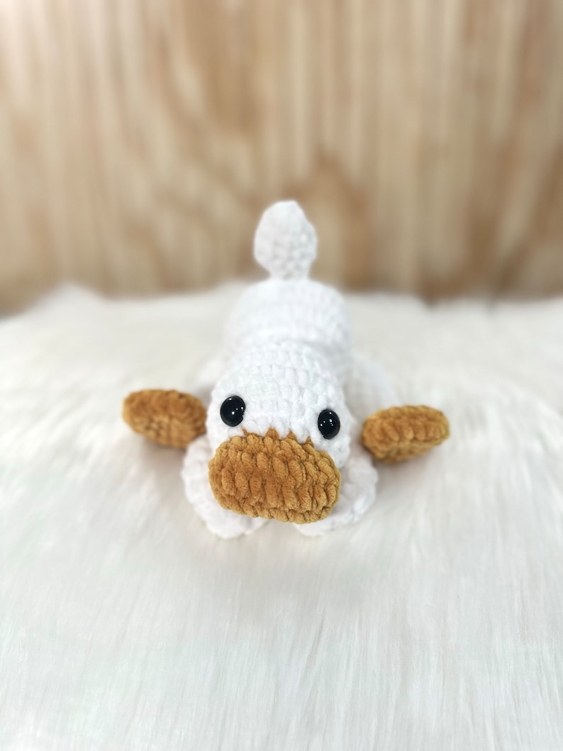 May include: A white crocheted duck plush toy with brown crocheted eyes and a brown crocheted beak. The duck is lying on a white fluffy surface.