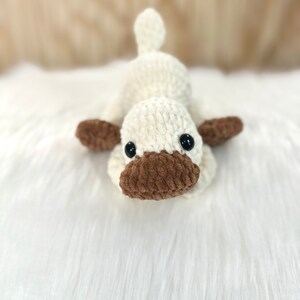 May include: A crocheted plush toy shaped like a duck. The duck is white with brown accents and black eyes. It is lying on a white fluffy surface.
