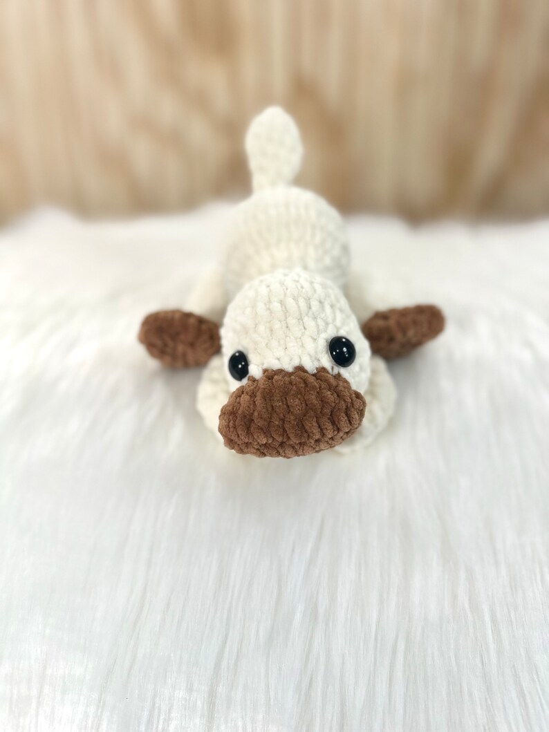 May include: A crocheted plush toy shaped like a duck. The duck is white with brown accents and black eyes. It is lying on a white fluffy surface.