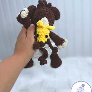 May include: A brown crocheted stuffed animal with a white diaper and a yellow crocheted stuffed animal with black eyes. The larger stuffed animal is holding the smaller one.