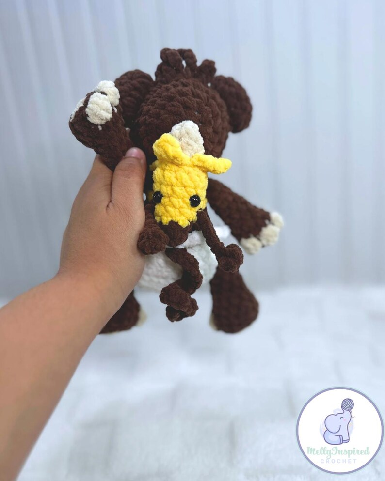 May include: A brown crocheted stuffed animal with a white diaper and a yellow crocheted stuffed animal with black eyes. The larger stuffed animal is holding the smaller one.