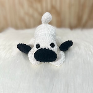May include: White crocheted dog toy with black crocheted eyes, nose, and ears. The toy is lying on a white fluffy surface.