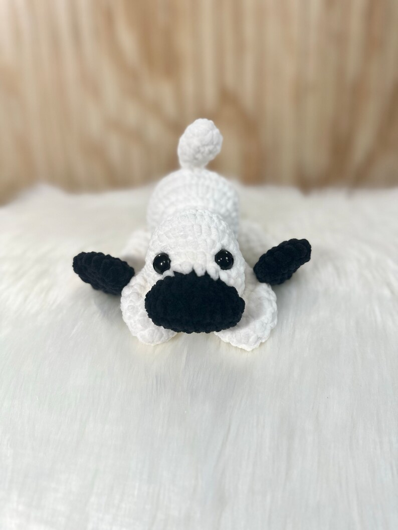 May include: White crocheted dog toy with black crocheted eyes, nose, and ears. The toy is lying on a white fluffy surface.
