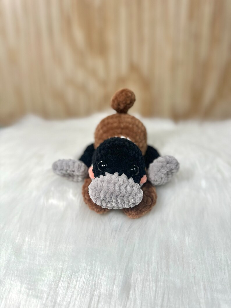 May include: A brown and black crocheted plush toy shaped like a platypus. The toy has a gray snout and black eyes.