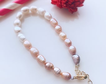 May include: A delicate pearl bracelet with a gradient of white, pink, and purple pearls. The bracelet has a gold clasp with a heart-shaped charm.