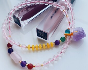May include: A pink rose quartz beaded bracelet with a charm featuring a purple amethyst stone, yellow beads, and green and blue stones. The bracelet is laying on top of a pink lipstick tube with the word "BURBERRY" printed on the side.