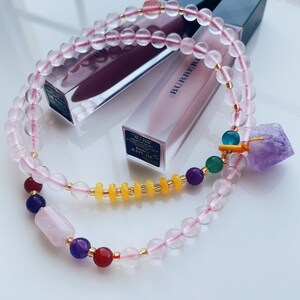 May include: A pink rose quartz beaded bracelet with a charm featuring a purple amethyst stone, yellow beads, and green and blue stones. The bracelet is laying on top of a pink lipstick tube with the word "BURBERRY" printed on the side.