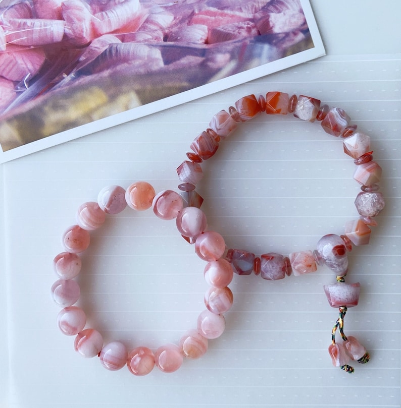 May include: Two pink agate bracelets with a tassel. One bracelet has round beads and the other has square beads.