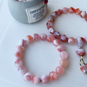 May include: Two bracelets made of pink and white agate beads. The bracelet on the left has round beads, while the bracelet on the right has faceted beads. Both bracelets have a red and white color scheme.