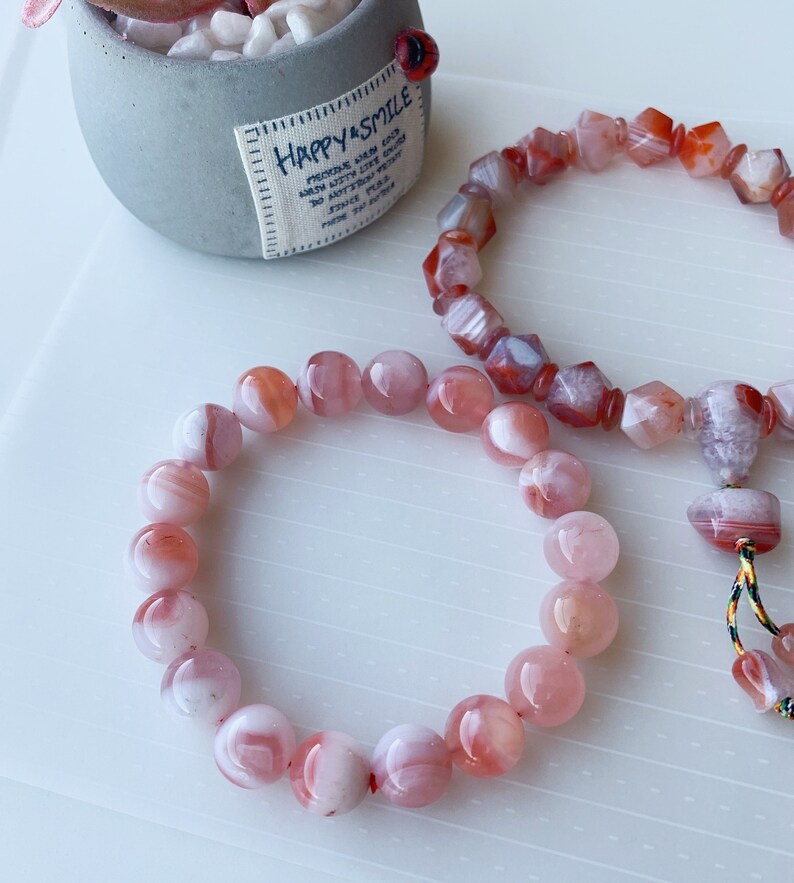 May include: Two bracelets made of pink and white agate beads. The bracelet on the left has round beads, while the bracelet on the right has faceted beads. Both bracelets have a red and white color scheme.