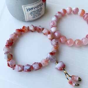 May include: Two bracelets made of pink and red agate beads. The bracelet on the left has faceted beads and a tassel. The bracelet on the right has round beads.