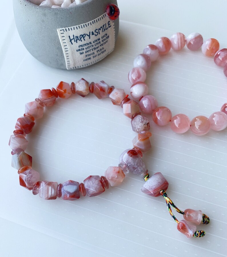 May include: Two bracelets made of pink and red agate beads. The bracelet on the left has faceted beads and a tassel. The bracelet on the right has round beads.