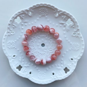 May include: A pink and white striped agate gemstone bracelet. The bracelet is made of round beads and is displayed on a white plate with a decorative design.