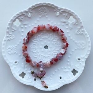 May include: A red and white agate gemstone bracelet with a tassel. The bracelet is made of square-shaped beads and is on a white background.