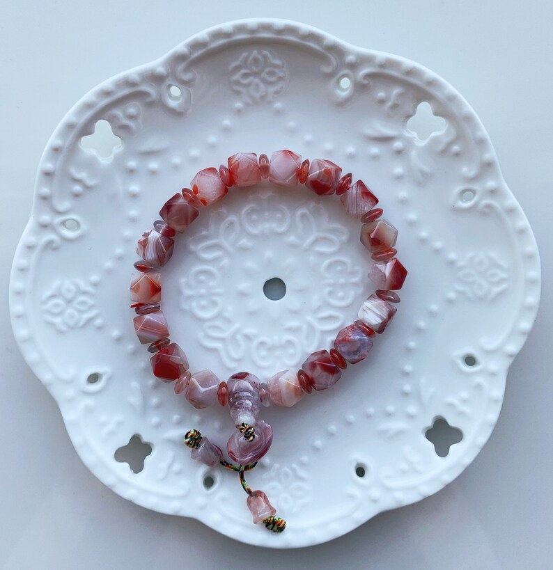 May include: A red and white agate gemstone bracelet with a tassel. The bracelet is made of square-shaped beads and is on a white background.