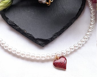 May include: A white pearl necklace with a red glitter heart pendant. The necklace is lying on a black heart-shaped slate with white flowers in the background.