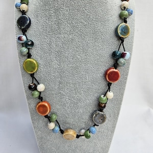 May include: A vibrant ceramic bead necklace on a grey mannequin. The necklace features a variety of round and oval beads in shades of orange, green, blue, and cream, strung on a black cord. The beads have a glossy finish.