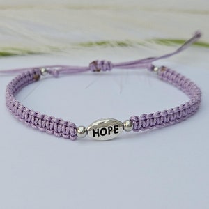Könnte beinhalten: Ein geflochtenes lila Armband mit einem silbernen Anhänger, auf dem "HOPE" steht.