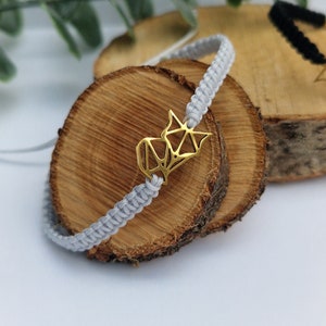 Puede incluir: Un brazalete trenzado gris con un dije geométrico dorado. El brazalete está sobre una superficie de madera.