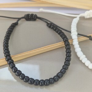 Puede incluir: Dos pulseras de la amistad, una negra y otra blanca, hechas con peque&ntilde;as cuentas redondas y atadas con un cord&oacute;n negro.
