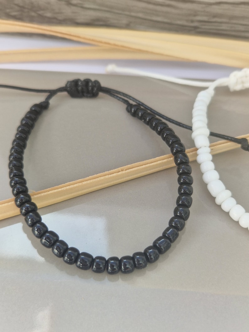 Puede incluir: Dos pulseras de la amistad, una negra y otra blanca, hechas con peque&ntilde;as cuentas redondas y atadas con un cord&oacute;n negro.
